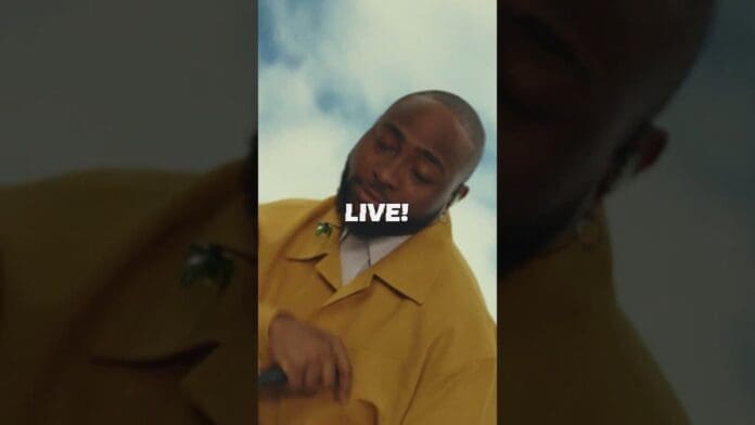 A person in a yellow coat holds a microphone and performs outdoors under a partly cloudy sky, channeling Lila Iké vibes, with the word "LIVE!" overlaid in white text.