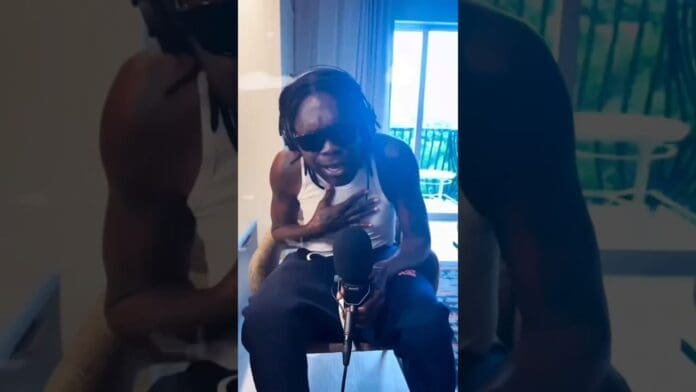 A person wearing sunglasses and headphones sits indoors, speaking into a microphone with one hand on their chest, passionately discussing Shatta Wale’s new music.