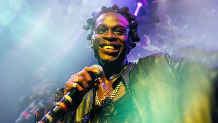A person with bantu knots smiles while holding a microphone on stage, wearing layered necklaces and a jacket, as colorful stage lights shine—just like a scene from a Fireboy DML lyrics video.