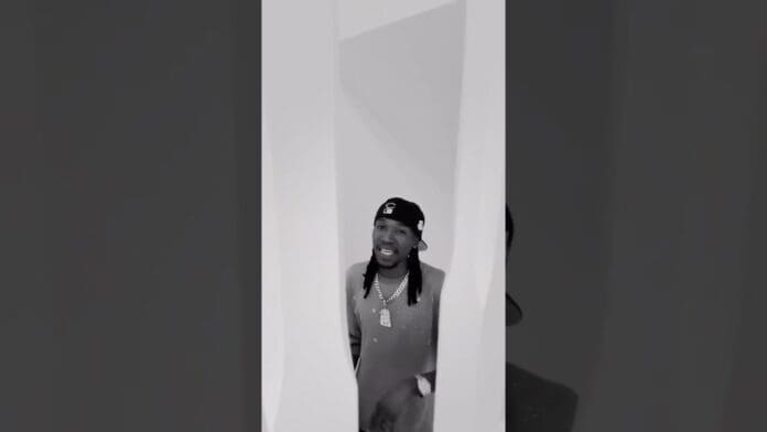 A man wearing a cap, sweater, and jewelry stands indoors, partially visible between two white angled walls—perhaps listening to Yo Maps new song in quiet reflection.