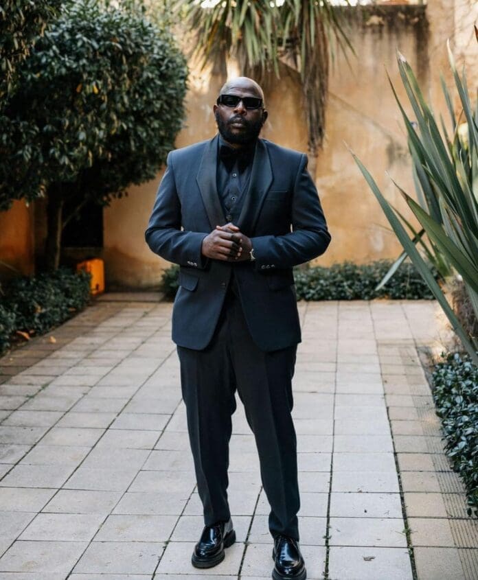 A man wearing a dark suit, bow tie, and sunglasses stands on a tiled outdoor path lined with greenery, exuding the cool style of afro house music artists like DJ Maphorisa.
