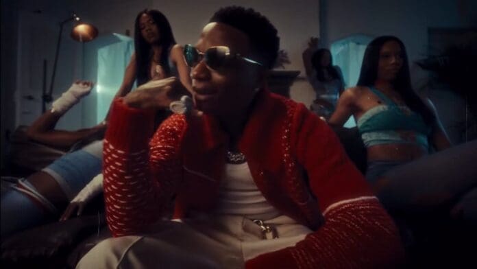 A man wearing sunglasses and a red sweater sits on a couch, surrounded by four women posing in a dimly lit room, as Amapiano beats inspired by DJ Maphorisa set the vibe.