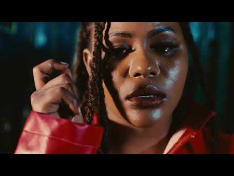 A person with braided hair and glossy makeup looks slightly downward, wearing a red jacket in a dimly lit setting—evoking the moody vibe of a Moyo Mjinga Official Video.