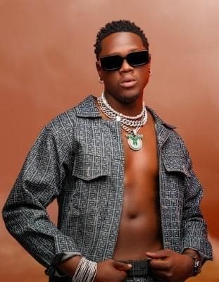 A man wearing sunglasses, layered necklaces, a patterned open jacket, and bracelets poses against a brown background, channeling Umthakathi Kush’s storytelling vibe with amapiano-inspired style.