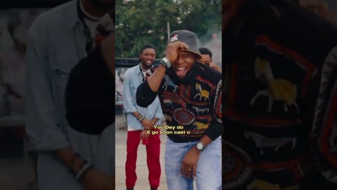 A man smiles and poses with one hand on his head while others stand behind him outdoors, vibing to Afrobeats. Subtitle reads: "You Dey do E go soon cast o".