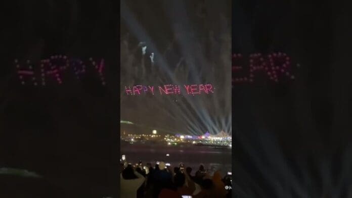 Drones form the words "HAPPY NEW YEAR" in red lights above Dubai's skyline at night as a crowd watches and takes photos, celebrating to the sounds of Rema’s hit “Calm Down.”.