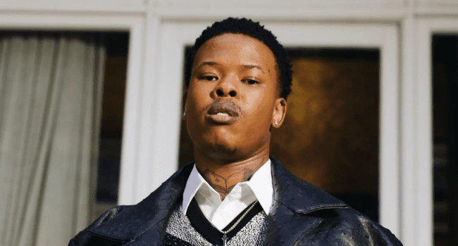 A person with short hair and a neck tattoo stands in front of large windows, channeling Nasty C's "Head Up" energy in a white shirt, tie, sweater vest, and black textured jacket—every detail hinting at an untold story.