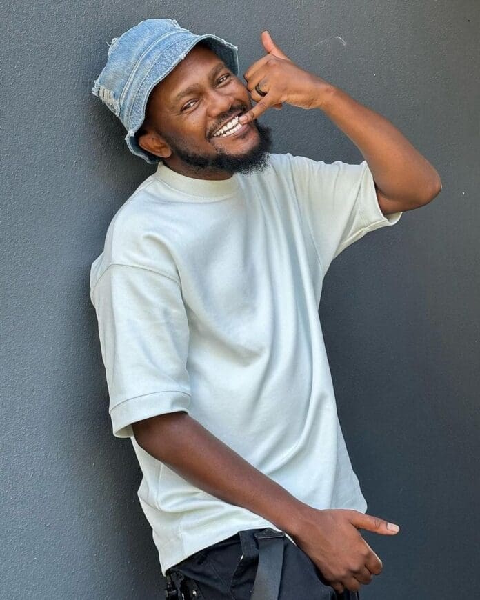 A man wearing a light bucket hat and t-shirt stands against a gray wall, smiling and making a hand gesture resembling a phone call, as if sharing his favorite South African music or discussing top albums from music albums 2016.