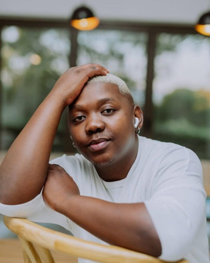 A person with short bleached hair wearing a white shirt and wireless earbud rests their head on their hand while seated indoors, lost in meaningful songs by Aymos from the Amapiano genre.
