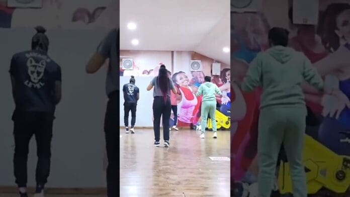 Four people are standing and facing a mirrored wall in a gym or dance studio, practicing dance moves with colorful posters in the background.