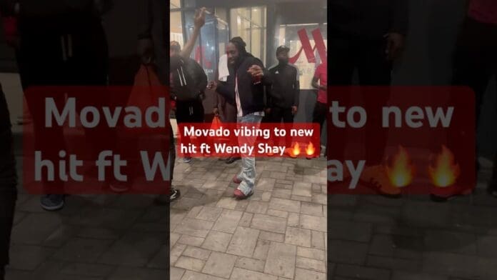 A man wearing a black jacket and red shoes dances dancehall-style on a sidewalk at night in front of a glass building, with several people nearby. Text overlay mentions "Movado vibing to new hit ft Wendy Shay.