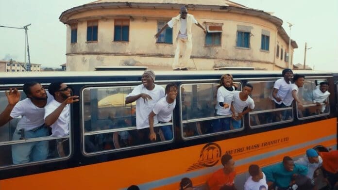 A group of people in white shirts lean out of the windows of an orange Metro Mass Transit bus, while one person stands on the bus roof—channeling LIL WIN’s energy—in front of a building, as if filming a lively Dance Video.
