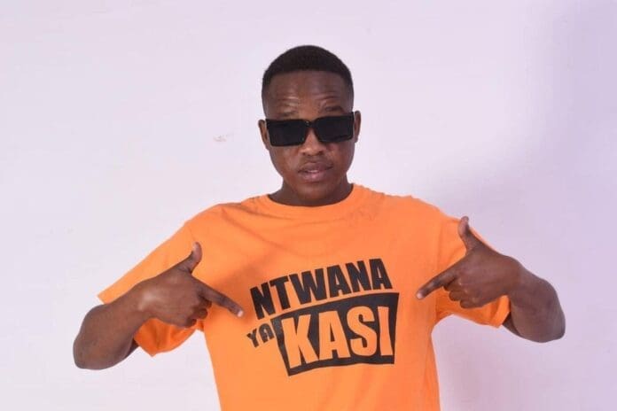 A person wearing dark sunglasses and an orange "NTWANA YA KASI" T-shirt stands against a plain light background, proudly pointing at the text—a true Ntwana Ya Kasi vibe inspired by Pampierstad and the amapiano culture.