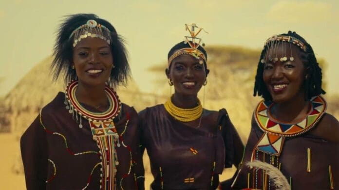 Three women wearing traditional attire and beaded jewelry stand outdoors, smiling, with thatched structures in the background—just like a scene from a Papalito & Kjack official music video.