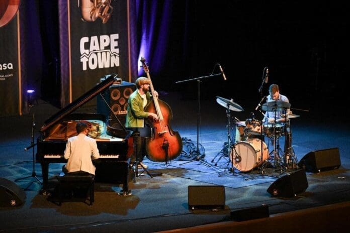 Three musicians perform on stage at the Cape Town Jazz Festival, one of South Africa music hubs, playing piano, double bass, and drums under vibrant stage lighting.
