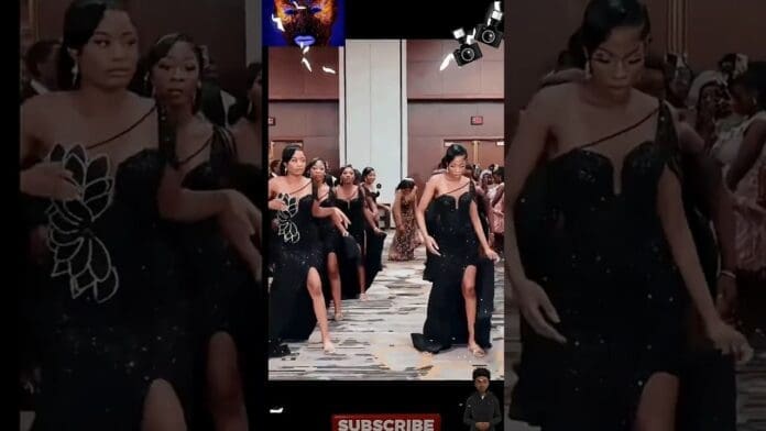 A group of women in black dresses with thigh slits are performing an amapiano dance in formation at an indoor event or party, captivating a crowd of onlookers.
