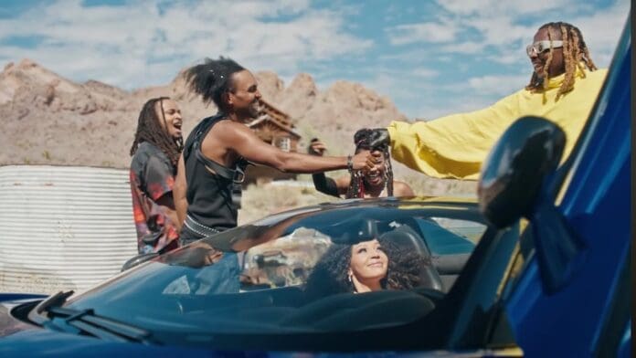 Four people stand outside in a desert setting, greeting each other near a blue car while Amapiano music plays softly; a woman sits inside the car and smiles.