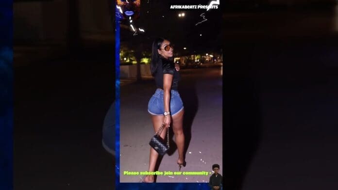 A woman wearing sunglasses, a black top, and denim shorts poses at night on a sidewalk, holding a black handbag. Text overlays are present at the top and bottom of the image, adding an afrobeat vibe to the stylish scene.