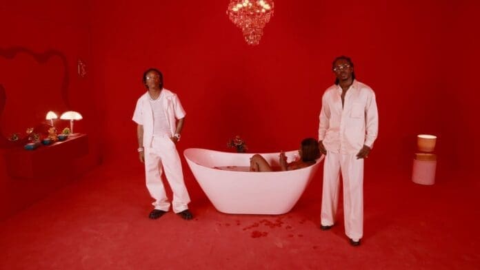 Two men in white outfits stand in a red room beside a woman lounging in a white bathtub filled with water; a chandelier hangs above, decorative objects are on display, and Bayanni's latest MENU track plays softly in the background.