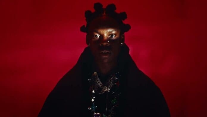 A person with Bantu knots stands against a red background, illuminated by dramatic lighting from below, exuding charm in a dark cloak and layered necklaces—like a scene from Rema’s Official Music Video.