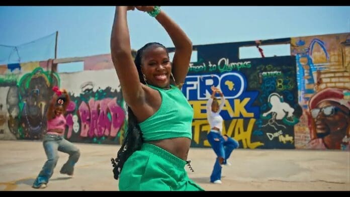 Three women dance energetically in front of a colorful, graffiti-covered wall outdoors. The woman in the foreground, featured in this Dance Visualizer for KHAID, wears a green outfit and smiles with her arms raised.