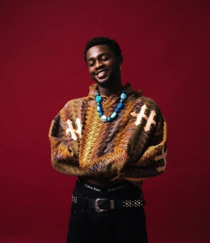 A person wearing a patterned brown sweater, blue beaded necklace, and black pants with a studded belt stands against a plain red background, smiling with arms folded at the WAIST.