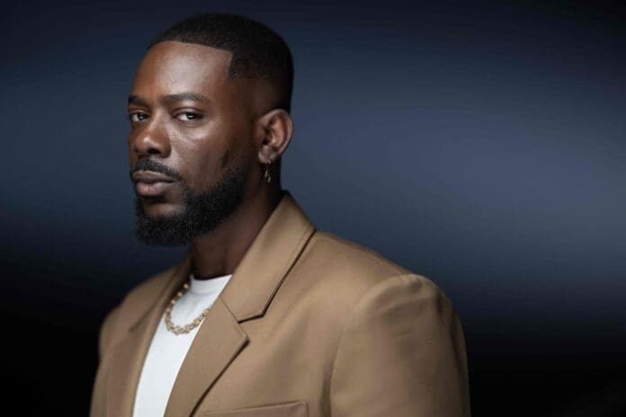 A man with a trimmed beard and short hair, wearing a tan blazer, white shirt, gold chain, and earring, channels Afropop style as he looks at the camera against a dark gradient background, reflecting his Nigerian roots.