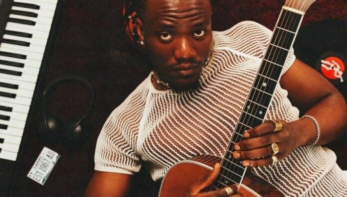 A man wearing a white mesh shirt lies on the floor holding an acoustic guitar, next to a keyboard and a vinyl record—capturing the relaxed vibe of Romantic Afropop Songs celebrated by OkayAfrica.