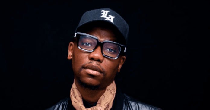 A man wearing glasses, a black "LA" cap, a brown patterned scarf, and a black jacket stands against a dark background, channeling Sowetan style and amapiano vibes.