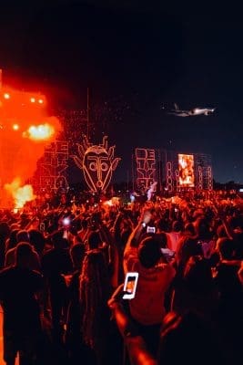 Crowd at a nighttime outdoor concert in Ghana with bright red lights, stage art, and a plane flying overhead; many attendees are holding up phones to record the electrifying Detty Rave event headlined by Mr Eazi.