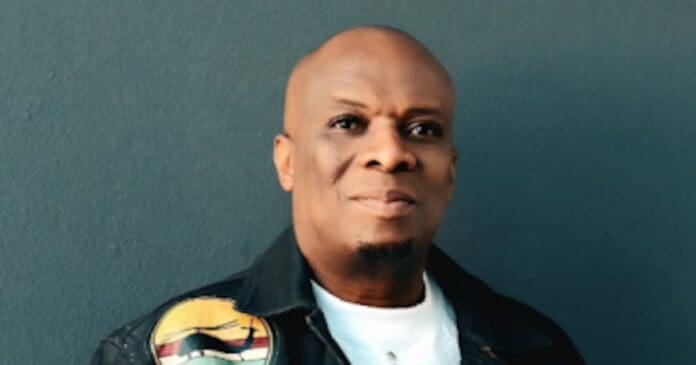 A bald man with medium-dark skin, resembling kwaito legend Thebe Lenyora, wears a black jacket with a colorful patch and a white shirt as he stands before a plain dark wall.