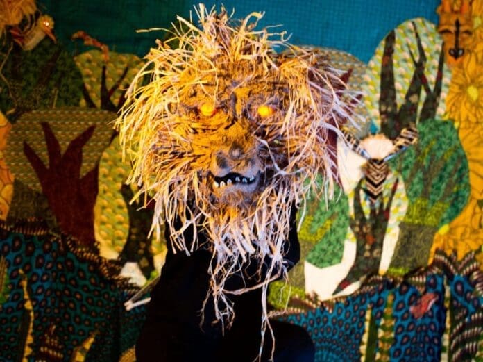 A person wearing a lion mask made of straw sits in front of a colorful, patterned fabric backdrop featuring trees, animals, and puppets.