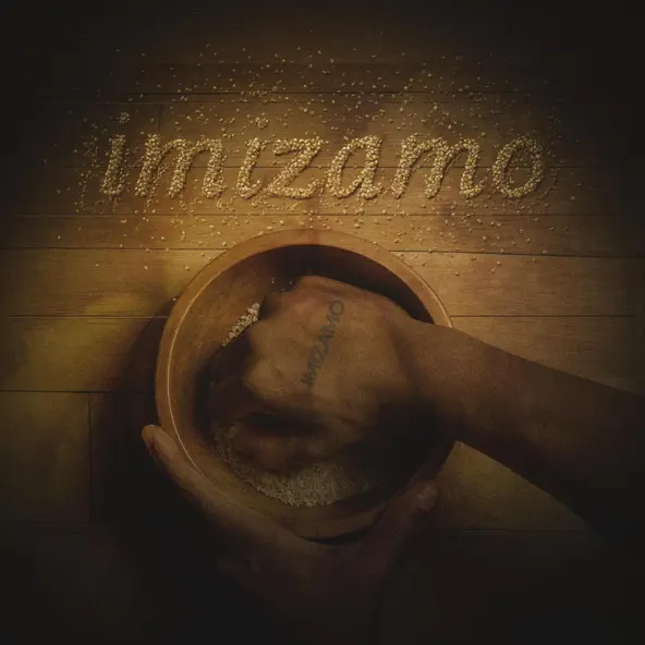 A hand mixes grains in a wooden bowl on a wooden surface. Above the bowl, the word "Imizamo" is spelled out using grains, inspired by the soulful sounds of Warren Justin.