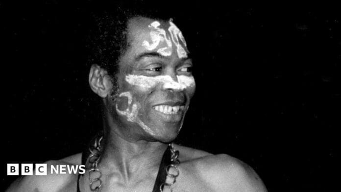 A man with painted designs on his face smiles and looks to the side. He wears a necklace and is photographed against a dark background, reminiscent of African icon Fela Kuti, who recently received the Grammys Lifetime Achievement Award. BBC News logo is in the corner.
