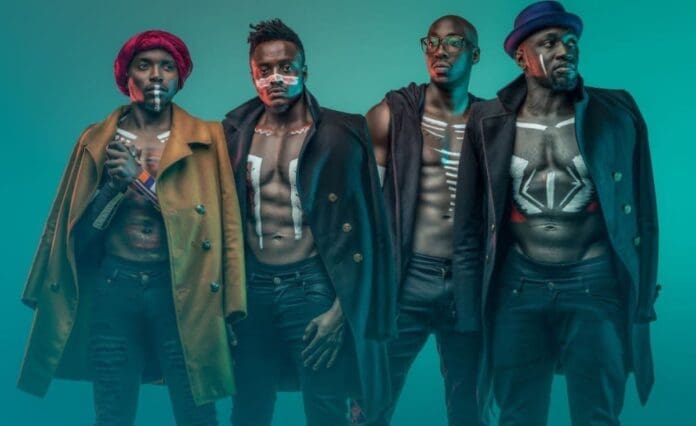 Four men of the Afro-Pop band Sauti Sol from Kenya wear open coats and painted tribal patterns on their chests, standing against a teal background with serious expressions and varied accessories.