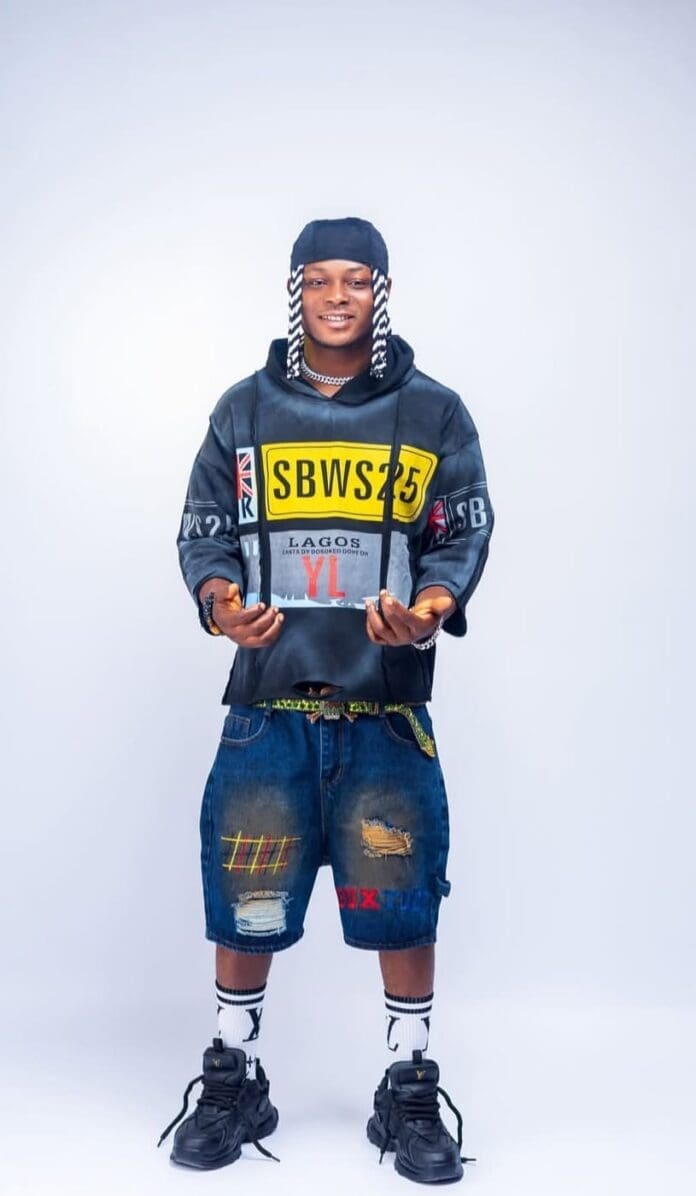 A person stands facing forward wearing a printed hoodie, ripped denim shorts, patterned socks, black sneakers, and a headscarf with long white braids against a plain background, channeling cool Lagos street style.