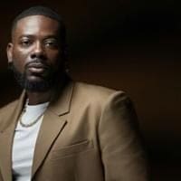 A man with a beard wearing a brown suit jacket, white shirt, and gold chain stands against a dark background, reflecting Adekunle Gold's Nigerian roots and Afropop style.