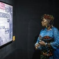 A person in colorful African traditional clothing looks at a framed newspaper display on a dark wall, highlighting Fela Kuti’s legacy and his Grammys Lifetime Achievement Award.