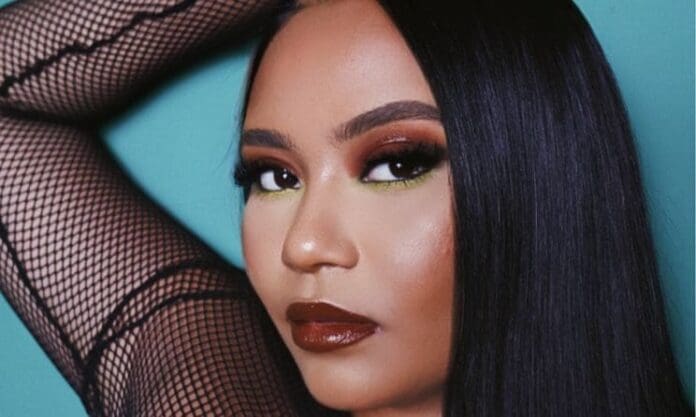 A woman with long straight black hair poses against a teal background, channeling MOLIY's bold Ghana style with fishnet sleeves, striking eyeshadow, green eyeliner, and dark brown lipstick.