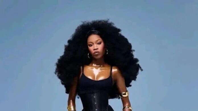 A woman with voluminous curly hair, wearing a black corset top, shiny gold arm cuffs, and hoop earrings, stands against a plain blue background—her confident pose echoes recent discussions on body-shaming controversy.