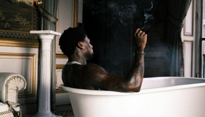 A man with tattoos sits in a white bathtub in a lavish room, holding a smoking item in his hand as ornate decor surrounds him—a scene reminiscent of July 2025 features on OkayAfrica celebrating Afrobeats songs.