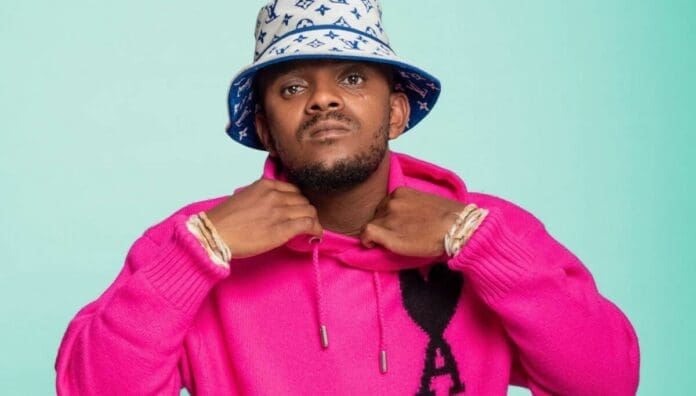 Man wearing a bright pink sweater and a patterned bucket hat stands against a plain light blue background, looking directly at the camera—giving off major July 2025 Amapiano songs vibes perfect for an OkayAfrica feature.