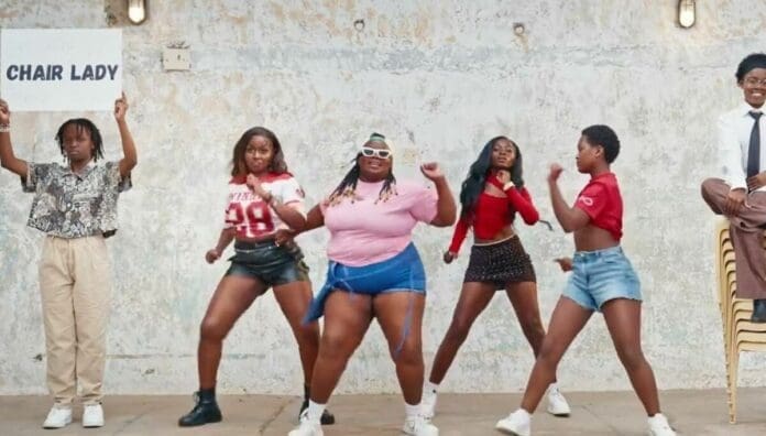 Five people dance in front of a concrete wall while one person sits on a stack of chairs at right; another holds a sign that reads "CHAIR LADY" on the left, capturing the spirit of July 2025 featured by OkayAfrica’s spotlight on East African Songs.