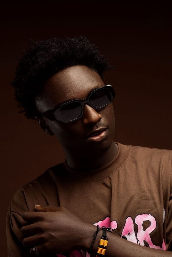 A person wearing dark sunglasses, a brown T-shirt with pink lettering, and a beaded bracelet poses against a dark background, channeling the confident style of an emerging Afropop artist.