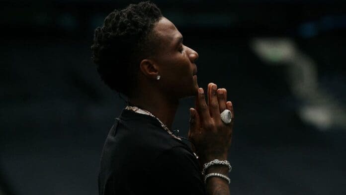 A person with short curly hair and jewelry clasps their hands together, looking upward, channeling the vibrant spirit of Afrobeats documentary style against a dark, blurred background.