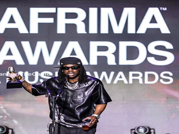 A person wearing sunglasses and a cap holds a trophy on stage at the AFRIMA Awards in Lagos, speaking into a microphone against a backdrop highlighting African music.