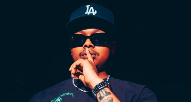A person wearing sunglasses, a black LA cap, and a watch holds a finger to their lips in a gesture for silence against a dark background—capturing the mysterious vibe of an A-Reece lead single.
