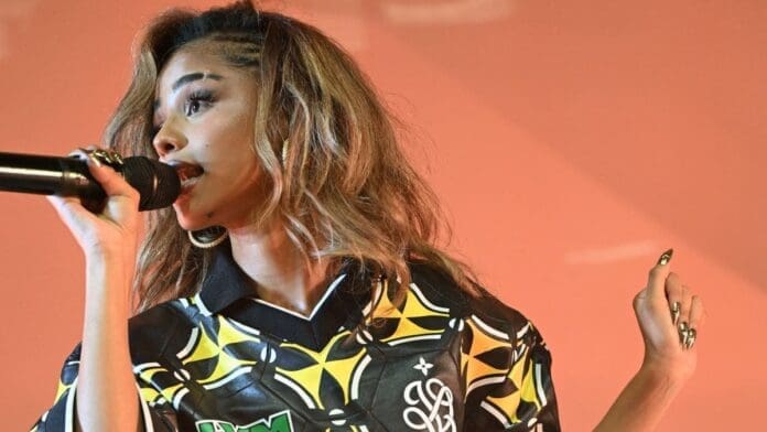 A woman with long wavy hair sings into a microphone on stage, wearing a patterned black and yellow shirt inspired by African music, set against a plain pink backdrop.