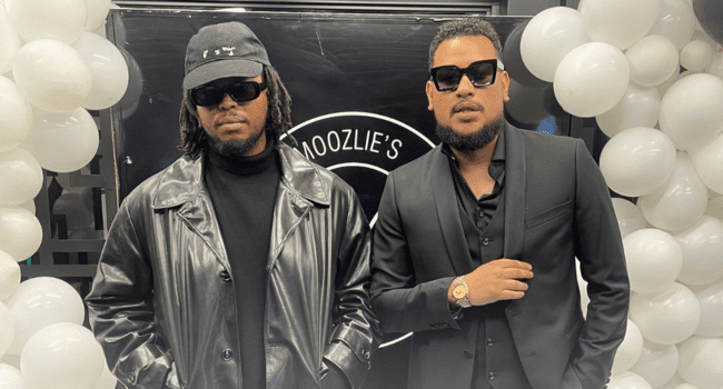 Two men wearing black outfits and sunglasses stand in front of a black and white balloon display and a sign that partially reads "Moozlie's," channeling the cool style seen in AKA and Anatii’s “10 Fingers” music video.