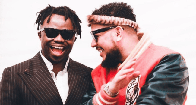 Two men are standing together, smiling and wearing sunglasses. One is dressed in a pinstripe suit, the other in a red and green jacket with a headband, posing for a tribute photo against a plain white background.